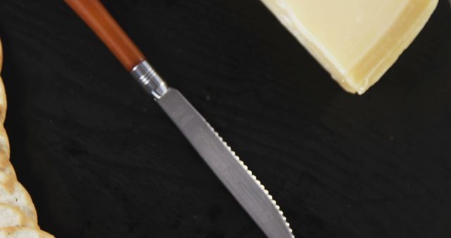 This close-up image features a serrated knife next to a block of butter and a cracker, set against a black background. It is ideal for use in food blogs, culinary websites, recipe illustrations, and advertising food-related products, emphasizing the elegance of simple, everyday kitchen items.