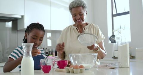 Senior woman teaching young girl baking in bright modern kitchen, sifting flour together