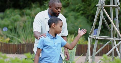 African american father guiding son in garden pointing at wooden trellis and plants