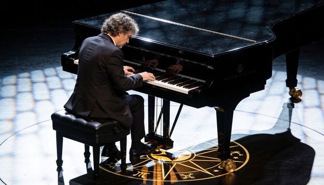 Pianist playing grand piano on spotlighted stage with decorative circular emblem in tuxedo