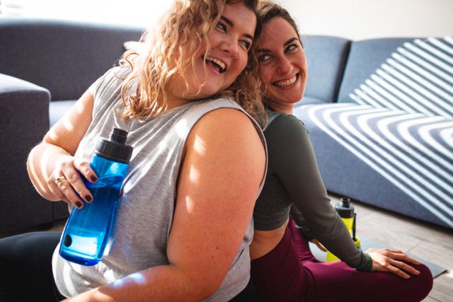 This image shows a joyful lesbian couple exercising together in their living room. They are sitting on the floor, leaning against each other, and holding water bottles. The scene is filled with natural sunlight, creating a warm and inviting atmosphere. This image can be used for promoting home fitness, LGBTQ inclusivity, healthy living, and domestic lifestyle content.