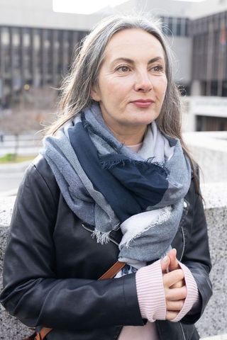 Mature woman leaning on concrete ledge in urban plaza wearing leather jacket and scarf