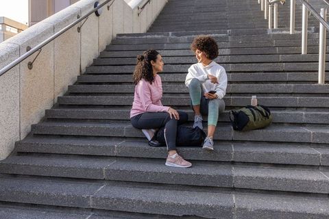 Young women sitting on campus stairs chatting after workout with backpacks and sneakers