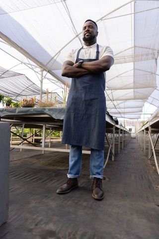 Confident african american nursery owner standing in greenhouse wearing denim apron