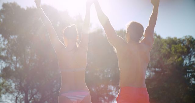 A young Caucasian woman and man are seen from behind, celebrating with their arms raised against a bright, sunny backdrop, with copy space. Their joyful gesture and casual summer attire suggest a sense of freedom and happiness during a warm outdoor activity.