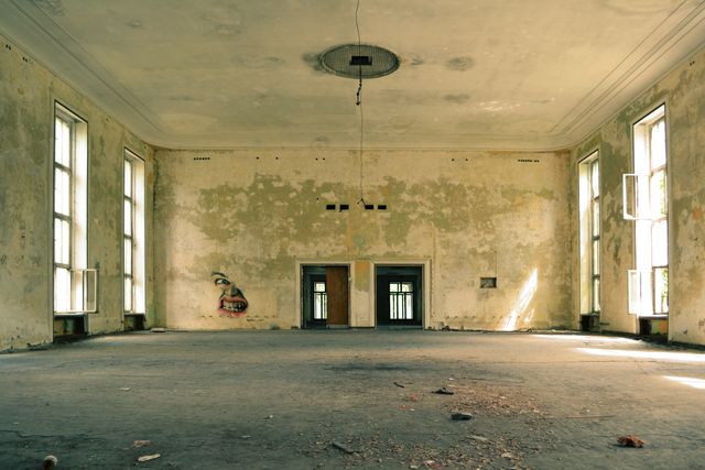 Spacious, empty room with large windows and aged, distressed walls. Ideal for concepts of urban decay, abandonment, or history. The hanging light fixture adds an element of nostalgia and neglect. Suitable for illustrating themes of bygone eras, desolation, or historical exploration.