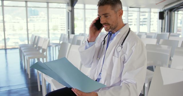 Caucasian doctor reviews a document in an office. He's multitasking in a bright medical office, consulting on the phone.