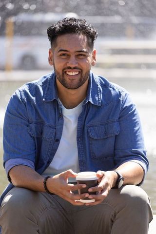 African american man smiling sitting and holding coffee cup by fountain in urban plaza