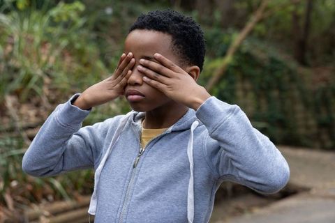 African american preteen covering eyes standing on park path wearing gray hoodie yellow tee