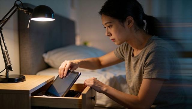 Woman sliding tablet into bedside drawer at night storing device in warm cozy bedroom