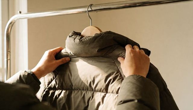 Adjusting olive puffer jacket collar on wooden hanger in sunlit wardrobe