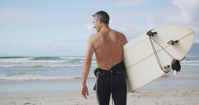 Senior man holding a surfboard while looking at ocean waves on a sunny beach. Ideal for use in promotions about active lifestyles, beach vacations, surfing culture, senior fitness, and adventure sports.