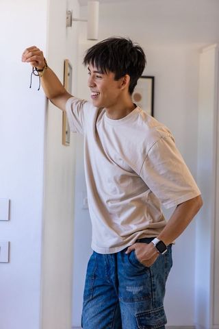 Smiling asian man leaning in hallway holding keychain wearing smartwatch and casual denim