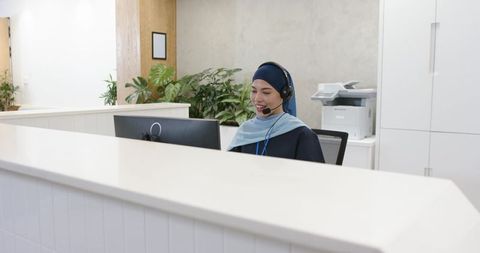 Smiling african american receptionist wearing headscarf speaking on headset at modern front desk