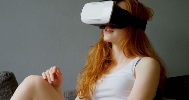Image shows woman with red hair using a virtual reality headset, engaging with technology in a casual home environment. Perfect for use in tech blogs, gaming websites, instructional materials, or advertisements promoting virtual reality equipment and entertainment.