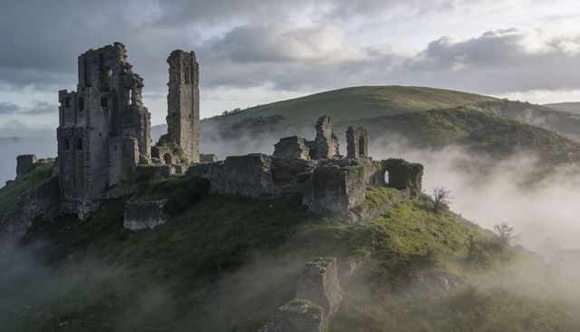 Ruined hilltop castle rising from morning mist over rolling green hills with ancient towers