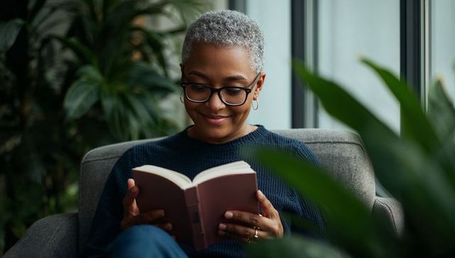 Serene senior woman reading in cozy armchair by window with plants and soft light