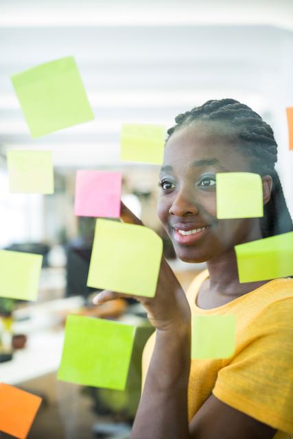 Young female graphic designer organizing colorful sticky notes on a glass wall in a modern office. Ideal for illustrating themes of planning, brainstorming, project management, creative work environment, and organizational skills. Suitable for use in articles, corporate presentations, and marketing materials related to creativity and professional strategy.