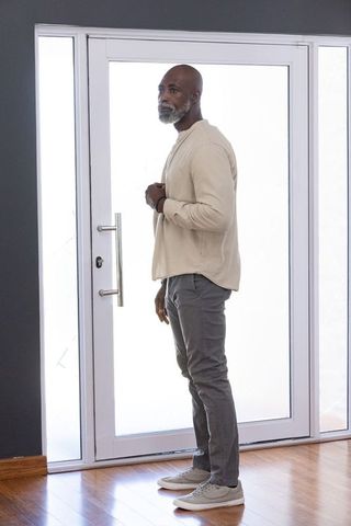 Senior african man standing by modern glass front door adjusting shirt, contemplative foyer
