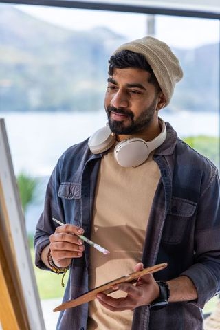 Contemplative male artist painting at easel with headphones in sunlit lakeside studio