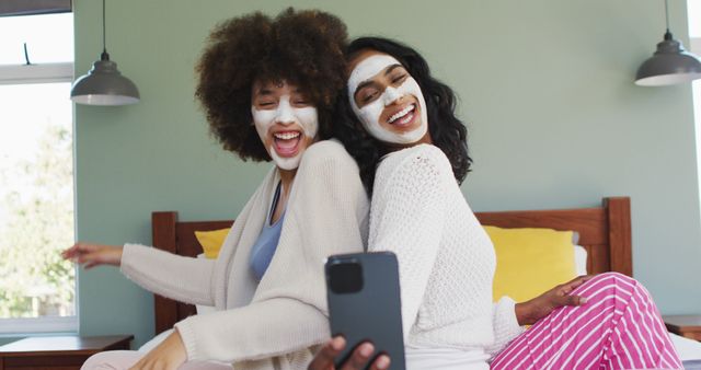 Two women apply face masks and sit back-to-back while enjoying the moment. They appear the capture the scene using a smartphone, documenting a light-hearted and enjoyable time together. This can be used for themes related to skincare routines, friendship bonding activities, relaxed self-care time, lifestyle brands, or beauty product advertisements.