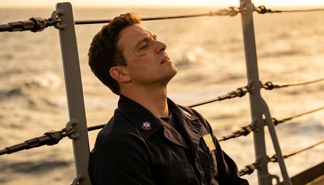Sailor sitting on ship rail closing eyes at sunset wearing navy coveralls and insignia