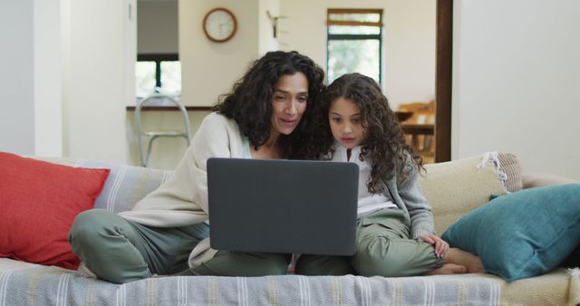 Mother and daughter sit together on a comfortable couch, using a laptop to access online learning resources in their home. They appear focused and engaged in their activity. This cohesive family moment highlights the opportunities for homeschooling, remote education, and the use of technology for learning in a familial setting. The image can be used for educational materials, advertisements for online learning platforms, or parenting articles.