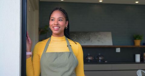 Smiling café barista leaning on doorway wearing olive-green apron and mustard top