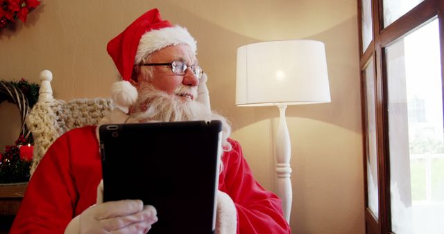 A senior Caucasian man dressed as Santa Claus is using a tablet, with copy space. His expression suggests he might be reading a digital list or preparing for the holiday season.