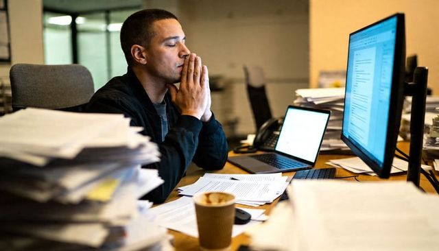 Stressed office worker pressing hands to face at cluttered desk with laptop and monitor