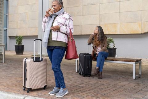 Mature multicultural women waiting with rolling luggage and coffee at transit station
