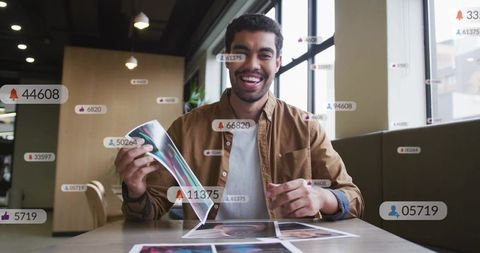 Smiling man holding glossy prints at cafe table with floating social media notifications