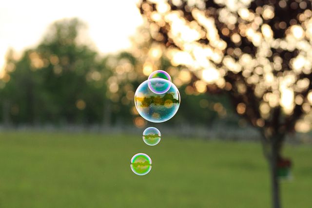 Colorful soap bubbles floating in green nature setting with trees and sunlight in background. Perfect for themes of childhood, play, outdoors, summer, and carefree moments. Ideal for websites, social media posts, and advertisements focusing on recreational activities or family fun.