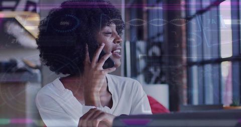 Speaking on smartphone at sunlit modern desk, woman working remotely with laptop