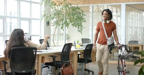 Young black professional walking bicycle into bright open office while greeting coworker