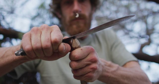 Man intensely focusing on sharpening a knife, holding the blade against a sharpening stone held in other hand outdoors. Ideal for depicting craftsmanship, skill, focus, outdoor survival, bushcraft, DIY tutorials, nature activities.