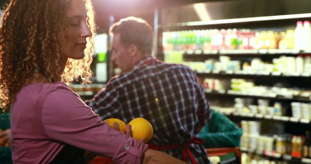 Woman with curly hair picking oranges in a grocery store while another shopper selects items nearby. Ideal for representations of everyday shopping, healthy lifestyle, fresh produce, supermarkets, and grocery shopping scenes.