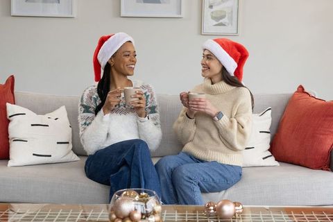 African american and asian women wearing santa hats holding mugs on sofa smiling chatting