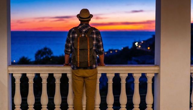 Solo traveler standing on balcony with backpack, gazing at vibrant coastal sunset horizon