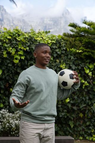 African american man holding soccer ball and gesturing in backyard garden with ivy