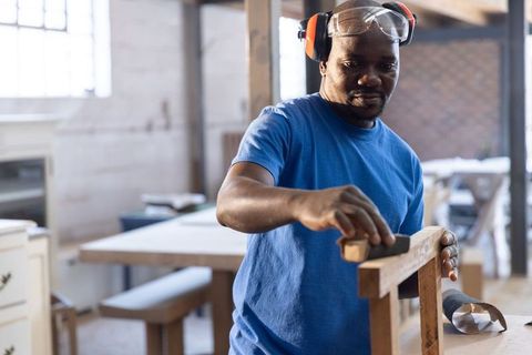 Mature african american carpenter sanding wooden chair frame wearing safety gear