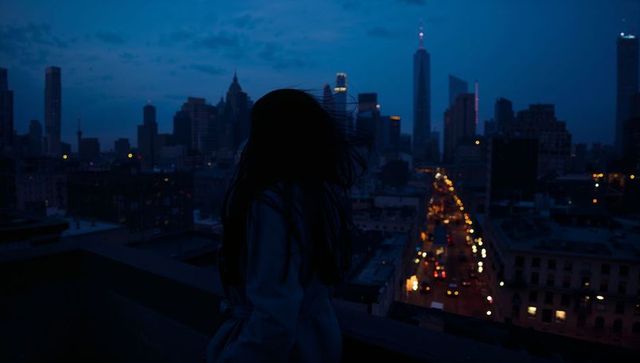 Silhouetted woman on rooftop at dusk overlooking lit city skyline, moody urban solitude