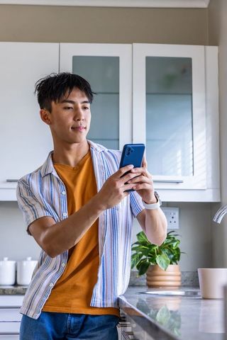Asian man leaning on modern kitchen counter holding smartphone and checking messages