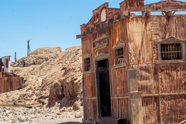 This image depicts an old, rusted building abandoned in a desert landscape under a clear blue sky. The weathered structure adds an eerie feel to the barren and desolate environment. Suitable for usage in themes related to history, desolation, abandonment, or to invoke a sense of forlorn eras or showcasing forgotten places. Ideal for travel blogs, adventure stories, or illustrative purposes in publications addressing historical sites or preservation.