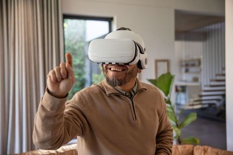Mature Asian man enjoying virtual reality at home wearing white VR headset and pointing