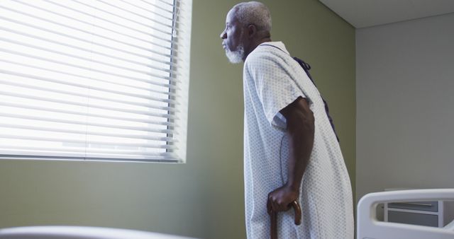African american male patient walking to window using cane in hospital room. medicine, health and healthcare services.