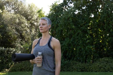 Mature woman holding yoga mat and shaker in garden promoting outdoor fitness and wellness