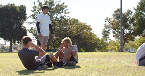 Diverse young men performing partner sit-ups on sunny park field with training cones