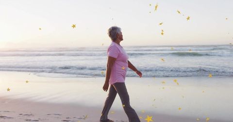 Senior woman walking along shoreline at sunrise with golden star confetti, peaceful stroll