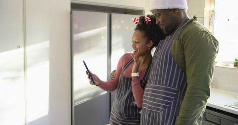 Couple Leaning Together Smiling at Phone Wearing Aprons in Bright Home Kitchen